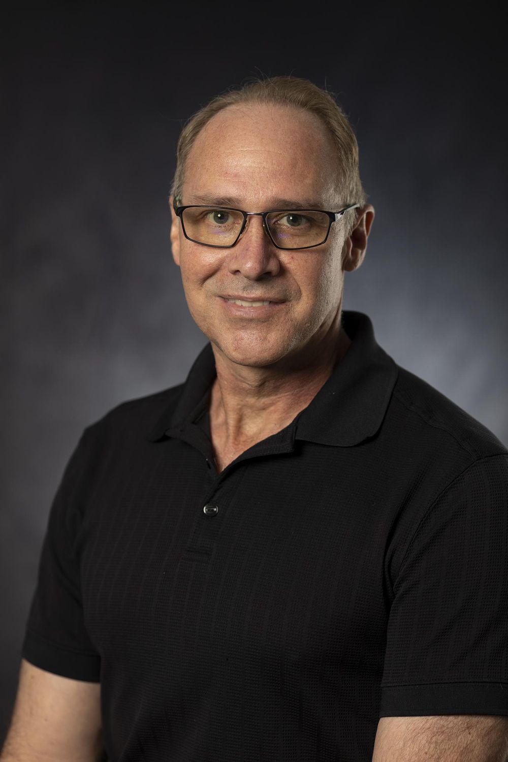 Business headshot of a smiling man with glasses in a black polo against a dark gray backdrop