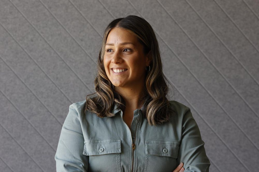 Natural-light portrait of a smiling woman in a sage button-up shirt against a textured gray wall