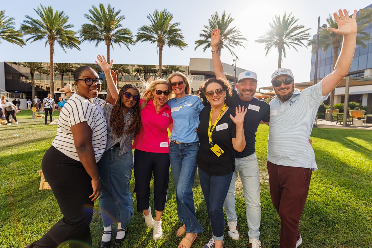 Colleagues cheering with raised arms at an outdoor company event on a sunlit lawn lined with palm trees
